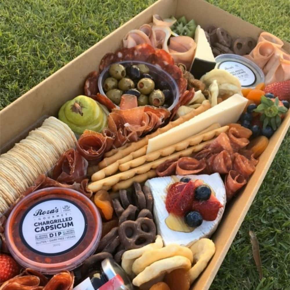 A low sided cardboard platter with an array of nibbles including breadsticks, ham, cracker and olives.