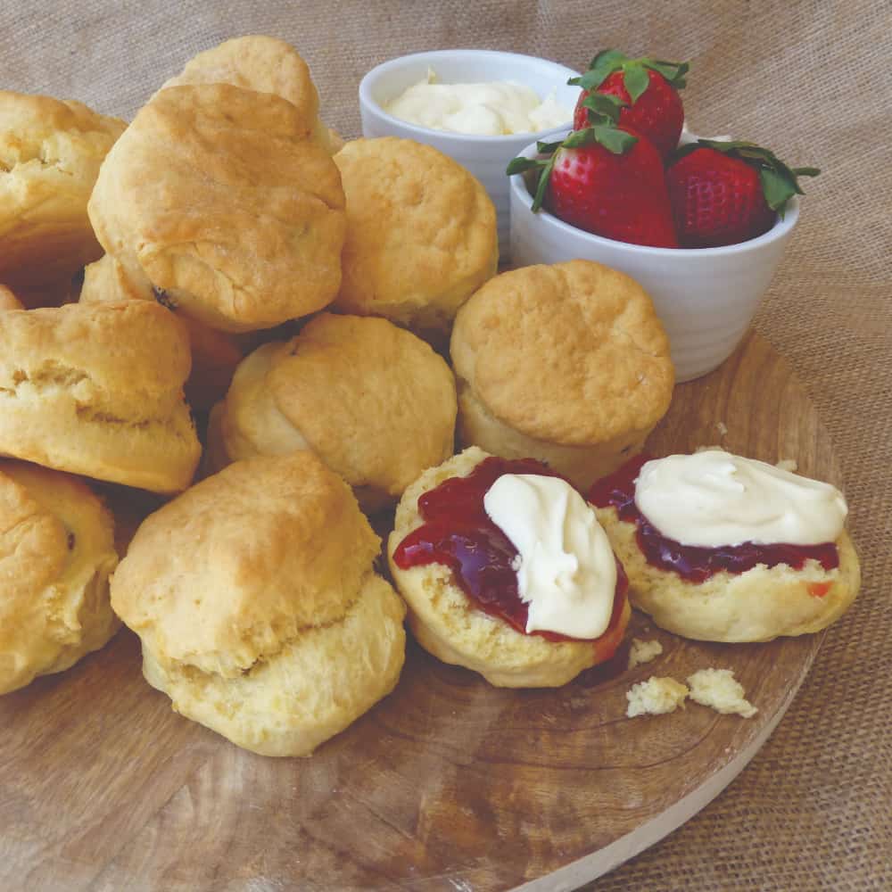 A wooden platter with a pile of freshly baked scones with jam and cream on a couple of them.